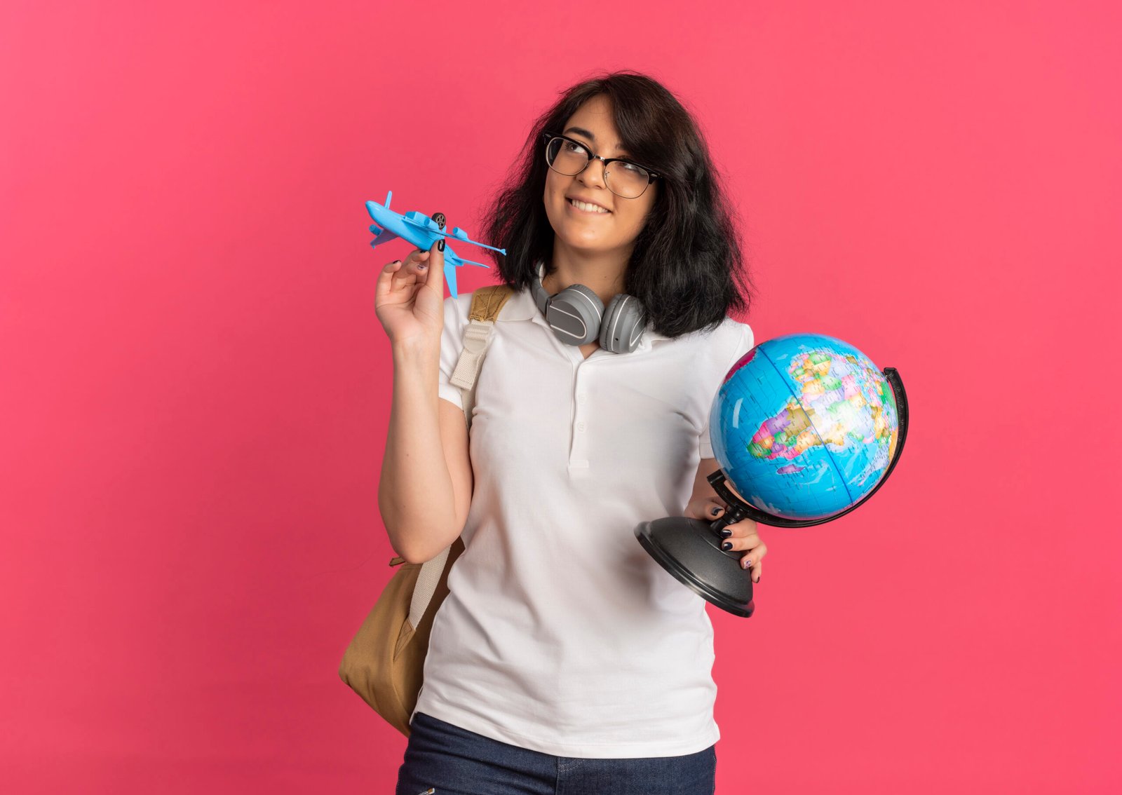 young pleased pretty caucasian schoolgirl with headphones neck wearing glasses back bag holds plane globe looking up pink with copy space scaled